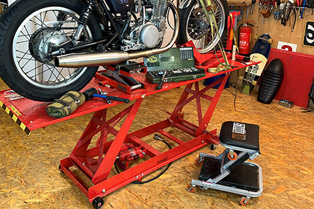 Auf einer Scherenhebebühne steht ein Motorrad, liegen diverse Handwerkzeuge, seitlich ein Rollhocker. 